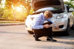 A man in a blue shirt inspects a damaged silver car's front bumper with an open hood, taking notes on a clipboard—an investigation process often supported by a Car Accident Claim Attorney to secure evidence