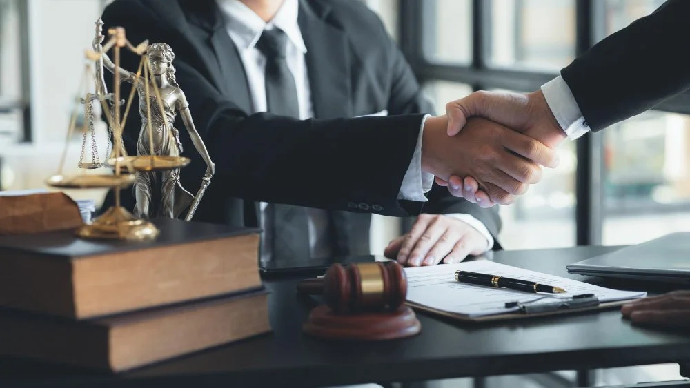 A Personal Injury Claim Lawyer shaking hands with a client across a desk set with a Lady Justice statue, law books, and a wooden gavel.
