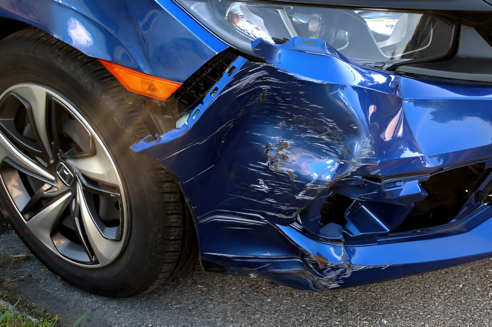 A detailed close-up of a damaged blue Honda car showing extensive scraping, crumpling, and fractured plastic on the front passenger-side bumper and fender. The scraped paint, broken grille sections, and damaged bumper trim are visible around the wheel, indicative of a car accident Hollywood Florida. The damage is concentrated near the lower fascia and headlight.